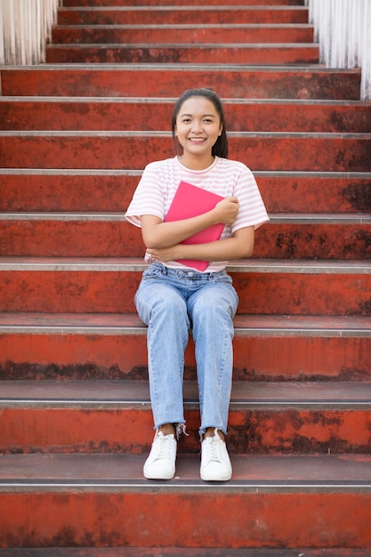 Niña de la escuela usa camisa rosa y jean sosteniendo y sosteniendo un libro rosa en las escaleras.