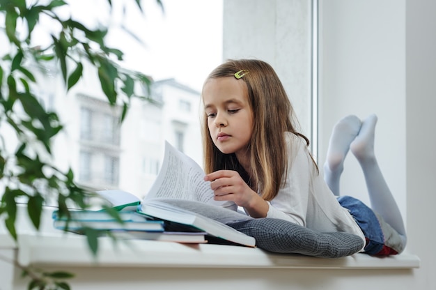 La niña está acostada en el alféizar de la ventana y lee un libro.