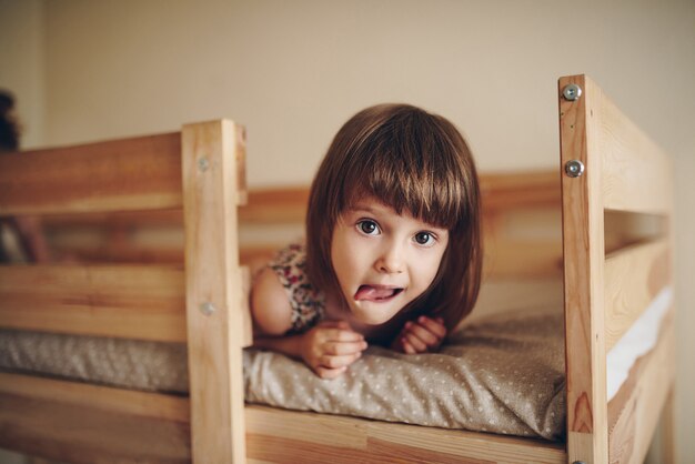 La niña está acostada en la cama, mirando a la cámara y mostrando la lengua.