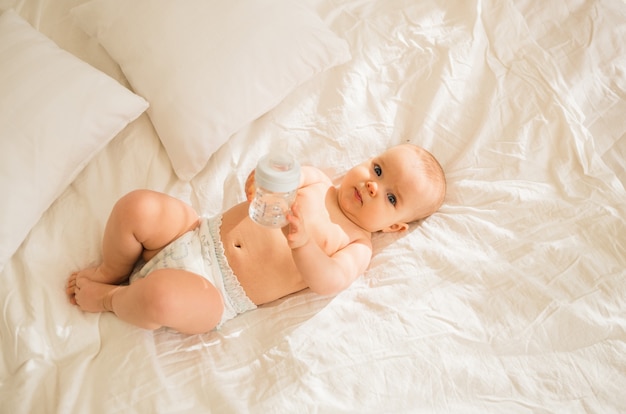 Una niña está acostada en un pañal con una botella de agua sobre la ropa de cama blanca en la cama