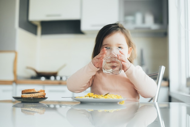 Una niña está en la cocina bebiendo agua hay un plato con una tortilla al lado