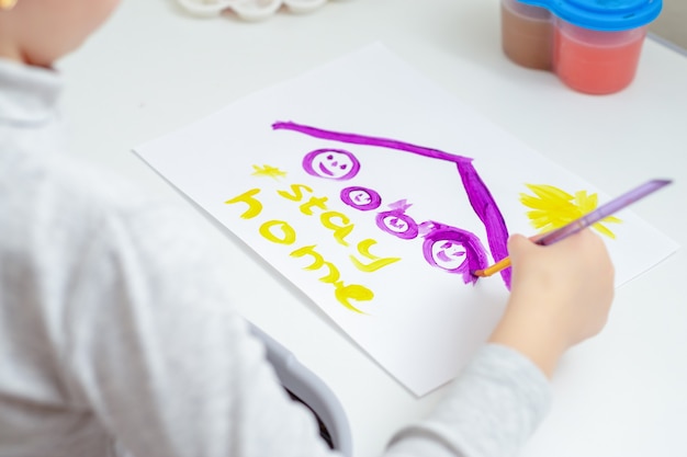 La niña está dibujando a la familia bajo un techo con las palabras Quédese en casa por acuarelas en una hoja de papel blanca. Concepto de estancia en casa.
