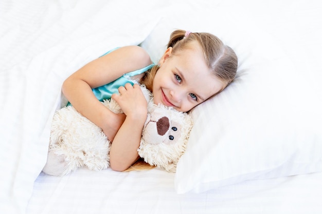 Una niña está durmiendo o se despertó por la mañana en la cama de su casa en una cama de algodón blanco abrazando un osito de peluche en sus manos y sonriendo dulcemente