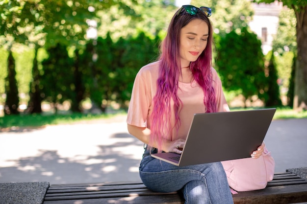 La niña está estudiando en el parque de la primavera sentada en el banco de madera y navegando en su computadora portátil