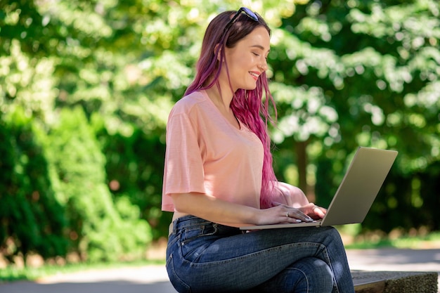 La niña está estudiando en el parque de la primavera sentada en el banco de madera y navegando en su computadora portátil