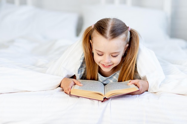Una niña está leyendo un libro en la cama de su casa en una cama de algodón blanco debajo de una manta y sonríe dulcemente