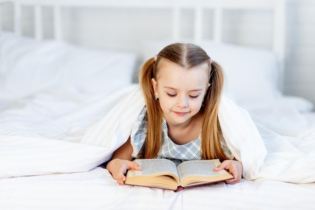 Una niña está leyendo un libro en la cama de su casa en una cama de algodón blanco debajo de una manta y sonríe dulcemente