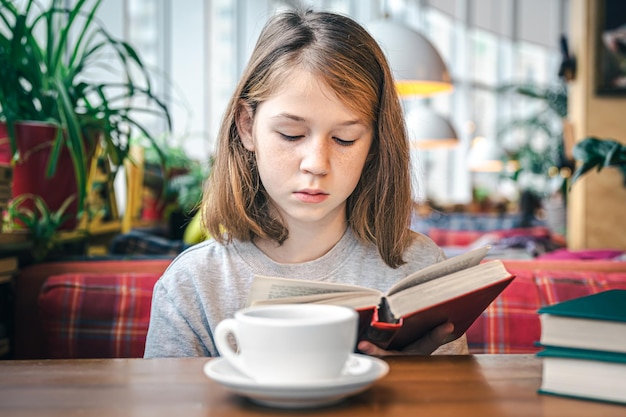 Una niña está leyendo un libro mientras está sentada en un café con una taza de té.