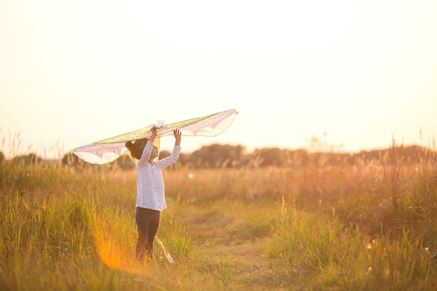 La niña está de pie con alas en el campo, aprendiendo a volar una cometa. Animación al aire libre en verano, naturaleza y aire puro. Infancia, libertad y descuido. Los sueños y la esperanza de los niños