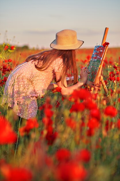 La niña está de pie en el campo de amapolas rojas y pinta sobre el lienzo colocado en un soporte de dibujo