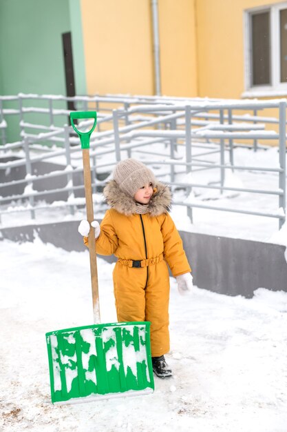 Una niña está de pie fuera de la casa en un día de invierno con una pala grande para quitar la nieve.