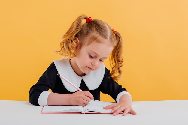 niña se está preparando para la escuela, escribe en el cuaderno, vestida con uniforme escolar sentado en la mesa blanca