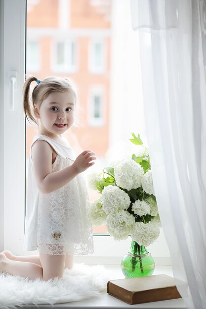 Una niña está sentada en el alféizar de la ventana. Un ramo de flores en un jarrón junto a la ventana y una niña oliendo flores. Una princesita con un vestido blanco con un ramo de flores blancas junto a la ventana.