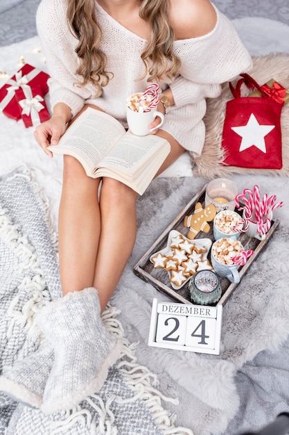 La niña está sentada en un ambiente navideño, tomando una bebida caliente y leyendo un libro.