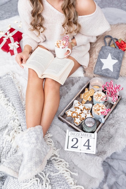 La niña está sentada en un ambiente navideño, tomando una bebida caliente y leyendo un libro.