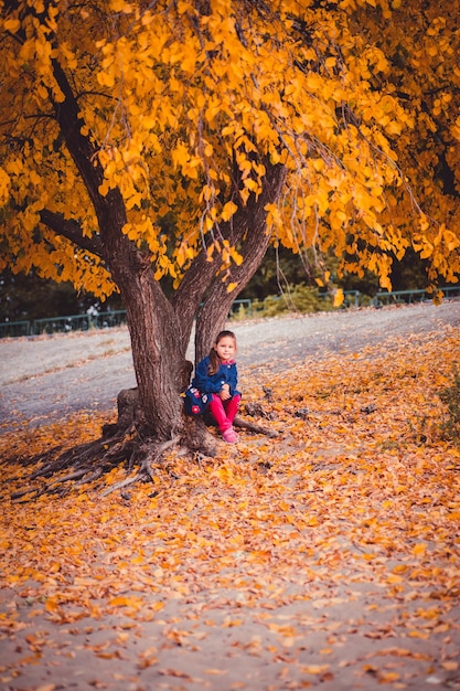 La niña está sentada bajo un árbol en el bosque y mirando a la cámara el niño en ropa de abrigo está descansando ...