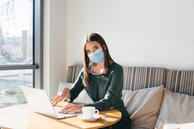 Una niña está sentada en un café con una máscara y trabajando en una computadora.