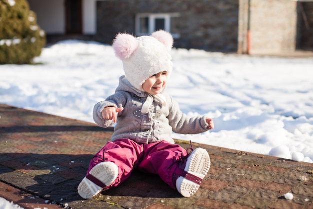 La niña está sentada en el camino en pantalones impermeables en un día frío y soleado de invierno