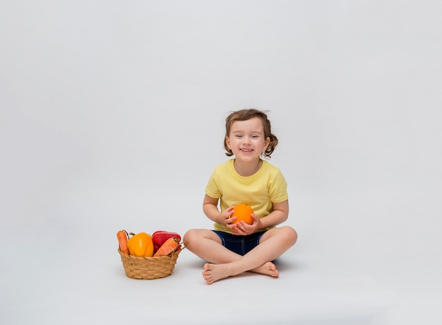 Una niña está sentada con una cesta de frutas y verduras en un espacio en blanco. Una linda chica con colas sonríe y sostiene una naranja en sus manos. Espacio libre.