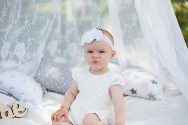 Una niña está sentada bajo un dosel sobre una manta blanca en un parque. Una niña linda mira hacia otro lado con un vestido blanco y una diadema en la naturaleza. La niña frunce el ceño