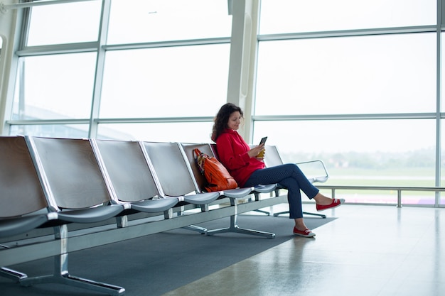 Una niña está sentada en una fila vacía de asientos frente a una gran vidriera en una terminal del aeropuerto, esperando un vuelo