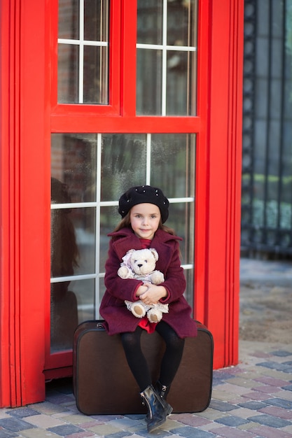 La niña está sentada en una maleta con un oso de peluche. Cabina telefónica roja de Londres. Primavera. Otoño. El viaje. Londres, Inglaterra. Primavera. Chica de escuela. Vacaciones escolares. Niña con boina y abrigo