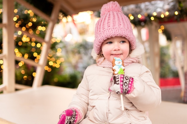 Una niña está sentada en el mercado de Navidad y está comiendo un pan de jengibre con un patrón de muñeco de nieve.