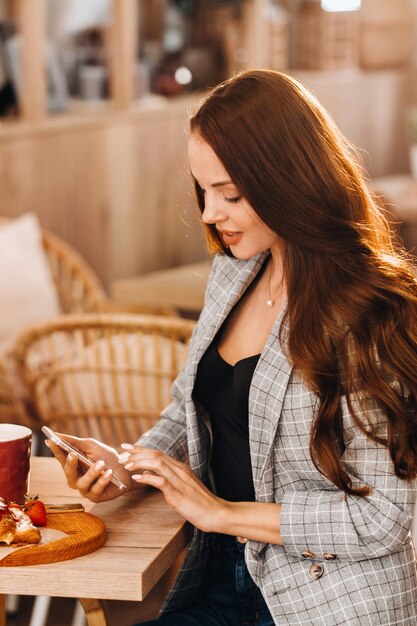 Una niña está sentada en una mesa y envía mensajes de texto en su teléfono inteligente en un café.Una niña está sentada en una cafetería con un teléfono.Escribe en el teléfono
