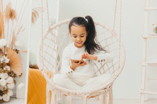 La niña está sentada y mirando su teléfono móvil, descansando sobre una silla colgante.