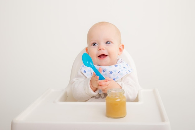 La niña está sentada en una silla alta sosteniendo una cuchara y comiendo puré de frutas