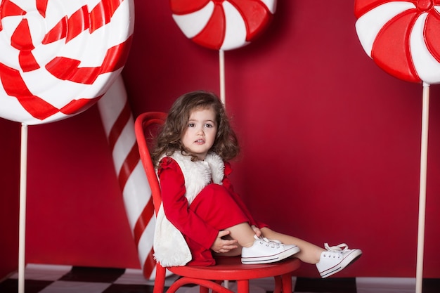 La niña está sentada en una silla con grandes caramelos de Navidad sobre fondo rojo.