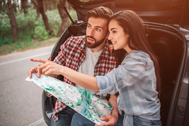 La niña está sentada con su novio en el baúl del auto y apunta hacia adelante