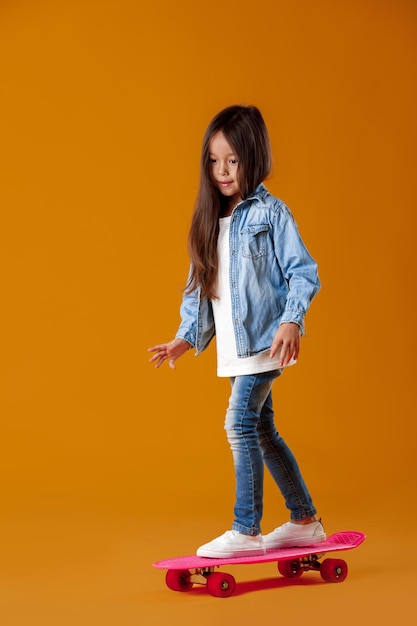 Niña con estilo niño con patineta en ropa de jeans sobre fondo naranja