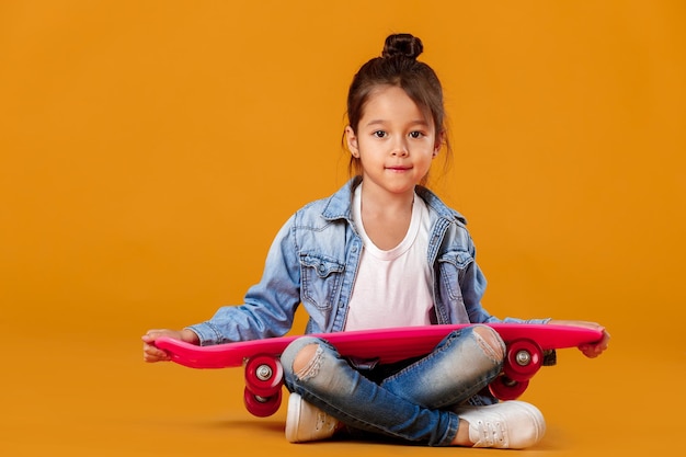 Niña con estilo niño con patineta en ropa de jeans sobre fondo naranja