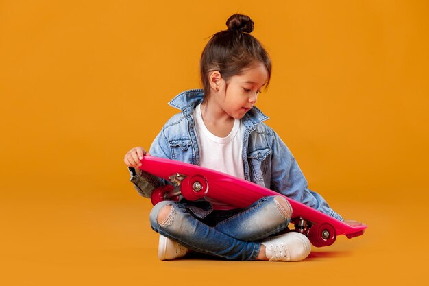 Niña con estilo niño con patineta en ropa de jeans sobre fondo naranja