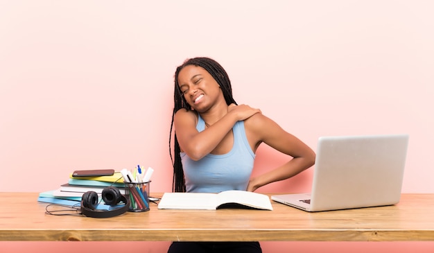 Niña estudiante afroamericana adolescente con cabello largo trenzado en su lugar de trabajo que sufre de dolor en el hombro