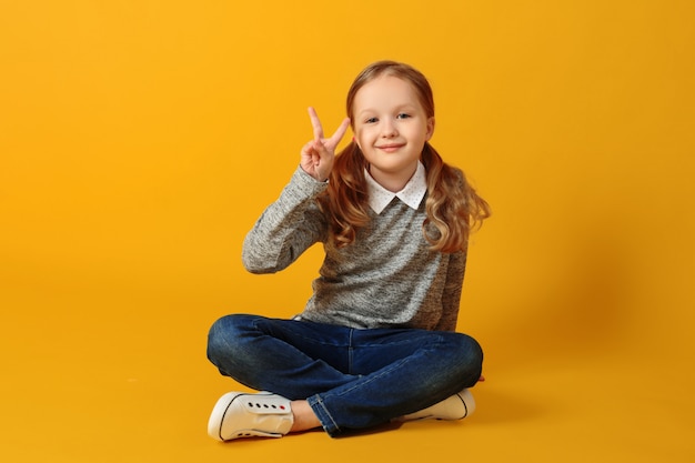 Niña estudiante está sentada en el suelo y mostrando un signo de victoria.