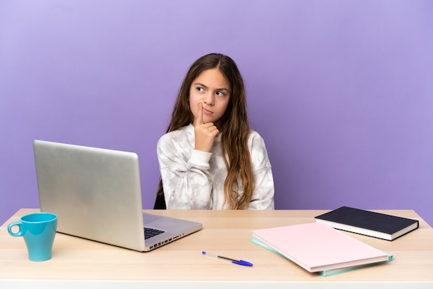 Niña estudiante en un lugar de trabajo con una computadora portátil aislada sobre fondo púrpura tener dudas mientras mira hacia arriba