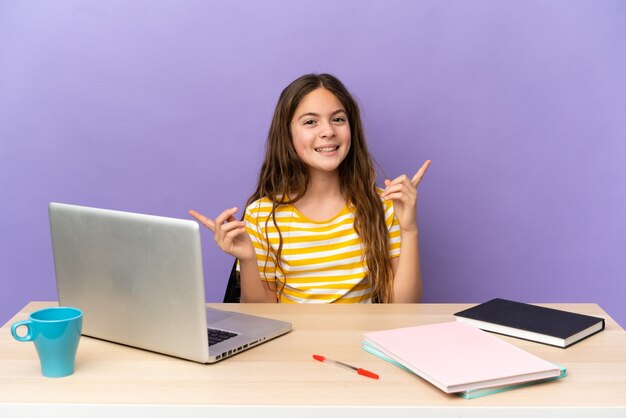 Niña estudiante en un lugar de trabajo con un ordenador portátil aislado sobre fondo púrpura dedo acusador a los laterales y feliz