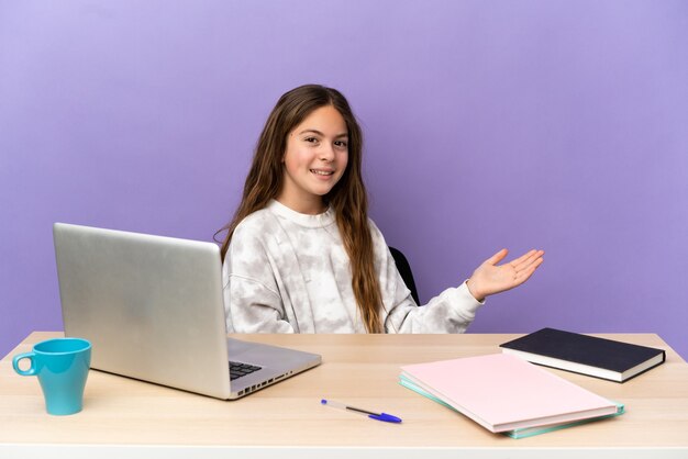 Niña estudiante en un lugar de trabajo con un portátil aislado sobre fondo púrpura extendiendo las manos hacia el lado para invitar a venir