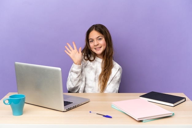 Niña estudiante en un lugar de trabajo con un portátil aislado sobre fondo púrpura saludando con la mano con expresión feliz