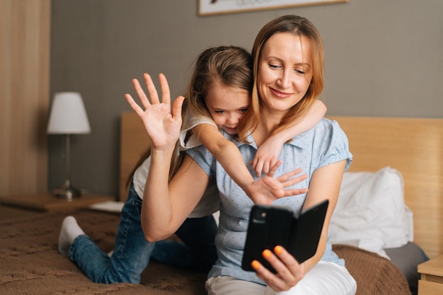 Niña feliz abrazando a una madre sonriente saludando agitando las manos durante una videollamada en línea usando un teléfono móvil, mirando una pantalla de teléfono inteligente. Concepto de actividad de ocio familiar en casa.