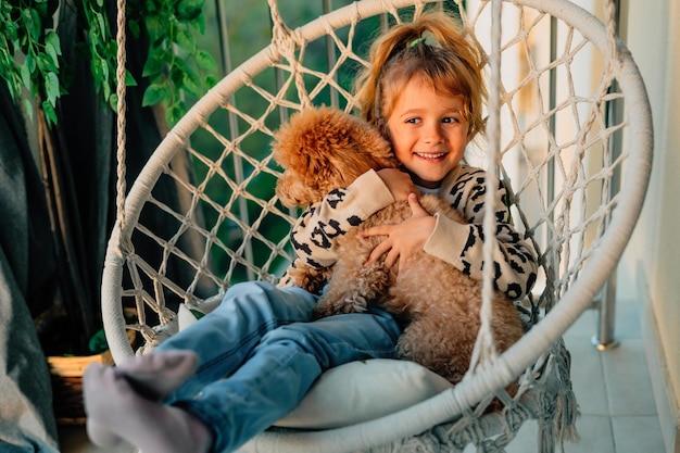 Niña feliz abrazando con una sonrisa a su perro caniche en casa en el balcón en primavera verano en una hamaca con flecos de algodón al atardecer