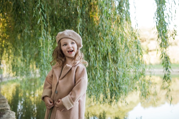 Foto una niña feliz con un abrigo beige y una boina está de pie cerca de un sauce