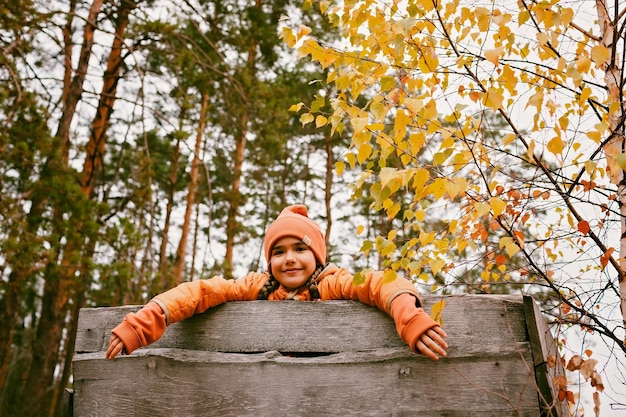 Niña feliz con abrigo naranja camina y juega en una casa de madera hecha a mano en el parque de otoño fin de semana activo