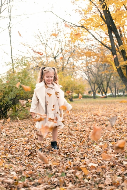 Niña feliz con abrigo y vestido arrojando hojas amarillas al aire en el parque de otoño