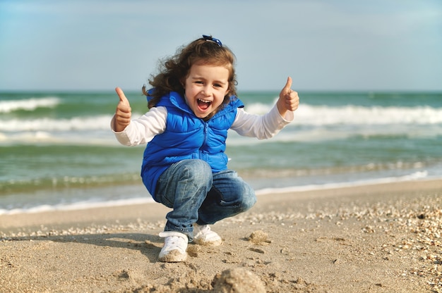 Niña feliz y alegre que muestra los pulgares para arriba mientras se pone en cuclillas en la playa. Día Internacional del Niño. Concepto de infancia feliz. Amor concepto de protección de la naturaleza y el planeta tierra