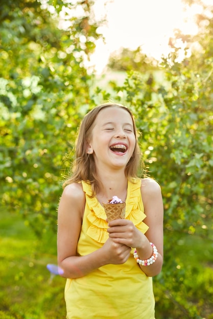Niña feliz con aparatos ortopédicos comiendo cono de helado italiano sonriendo mientras descansa en el parque el día de verano