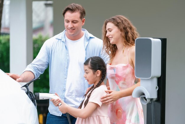 Una niña feliz aprende sobre la sostenibilidad ecológica y energética mientras ayuda a su familia a recargar el vehículo eléctrico desde su casa. Estación de carga de vehículos eléctricos. Coche eléctrico y concepto familiar moderno Synchronos