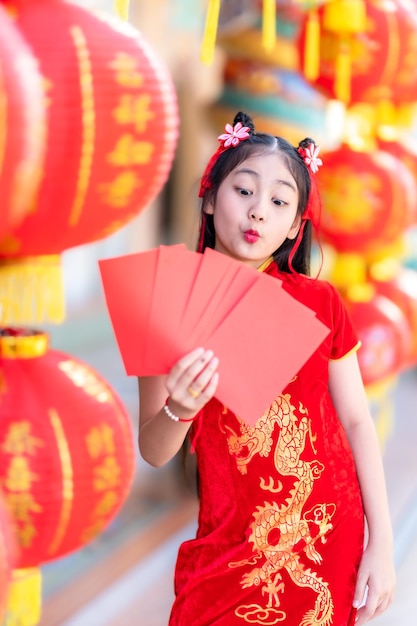 Niña feliz asiática con decoración de cheongsam chino tradicional rojo sosteniendo sobres rojos en la mano y farolillos con el texto chino Bendiciones escrito en él Es una bendición de la fortuna para los chinos